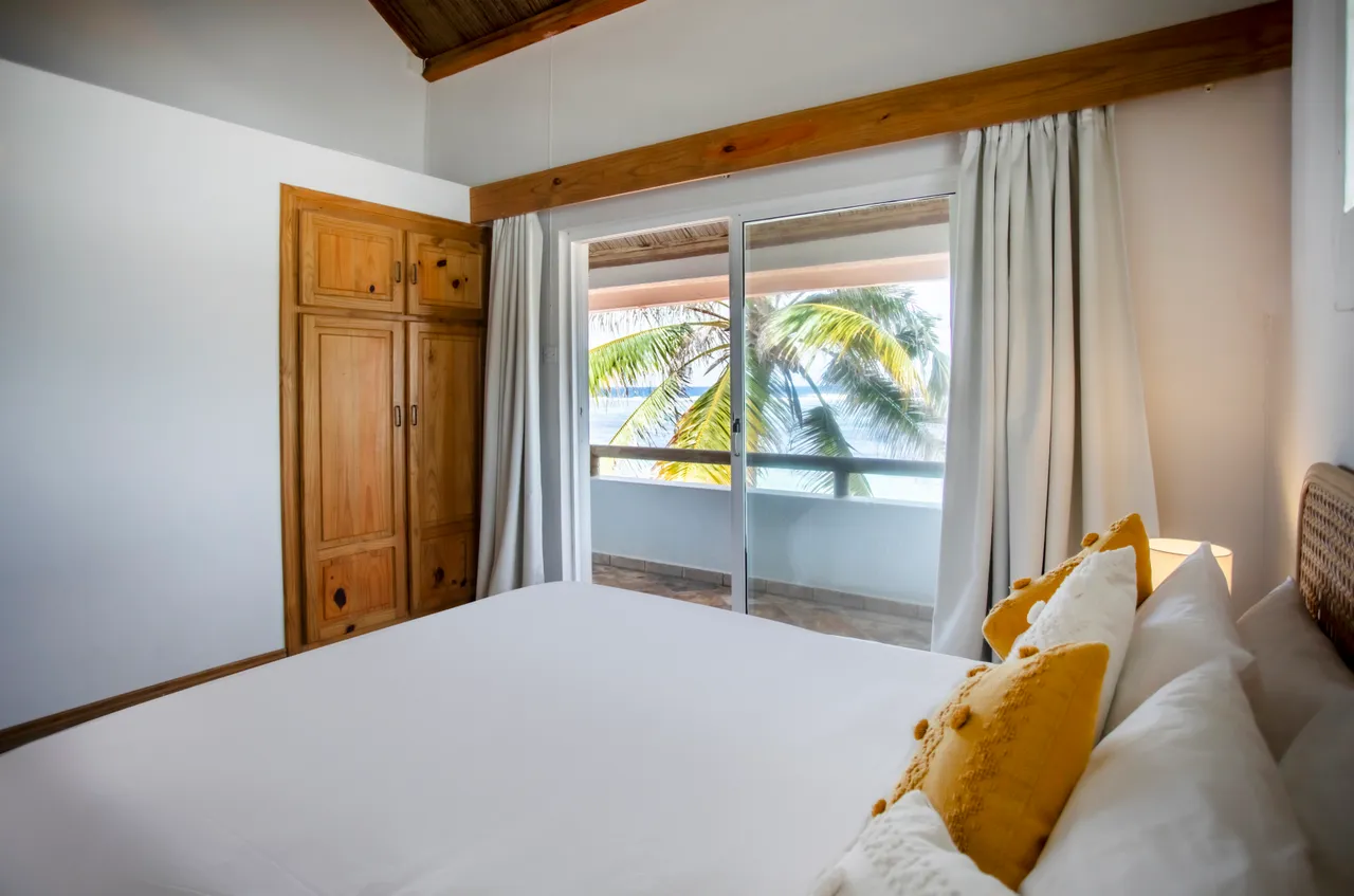 Chambre avec lit double et rangement en bois. Grandes fenêtres offrant vue sur la mer. Décor simple avec textiles clairs et lumineux.