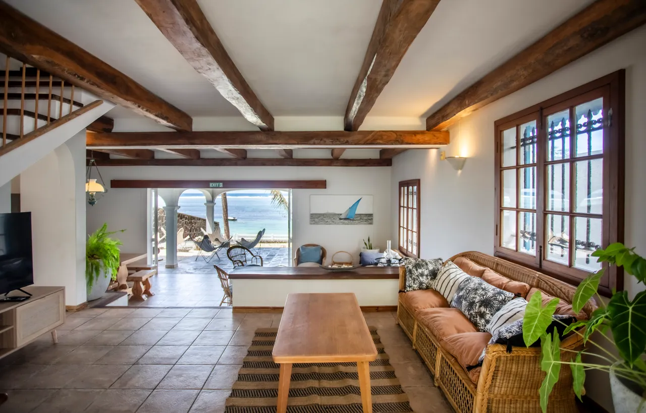 Lumière naturelle grâce aux grandes fenêtres, ouvrant sur la mer. Espace de vie avec canapés en rotin et table en bois. Plafond avec poutres apparentes, créant une ambiance chaleureuse. Accès direct à la terrasse extérieure, offrant une vue dégagée.