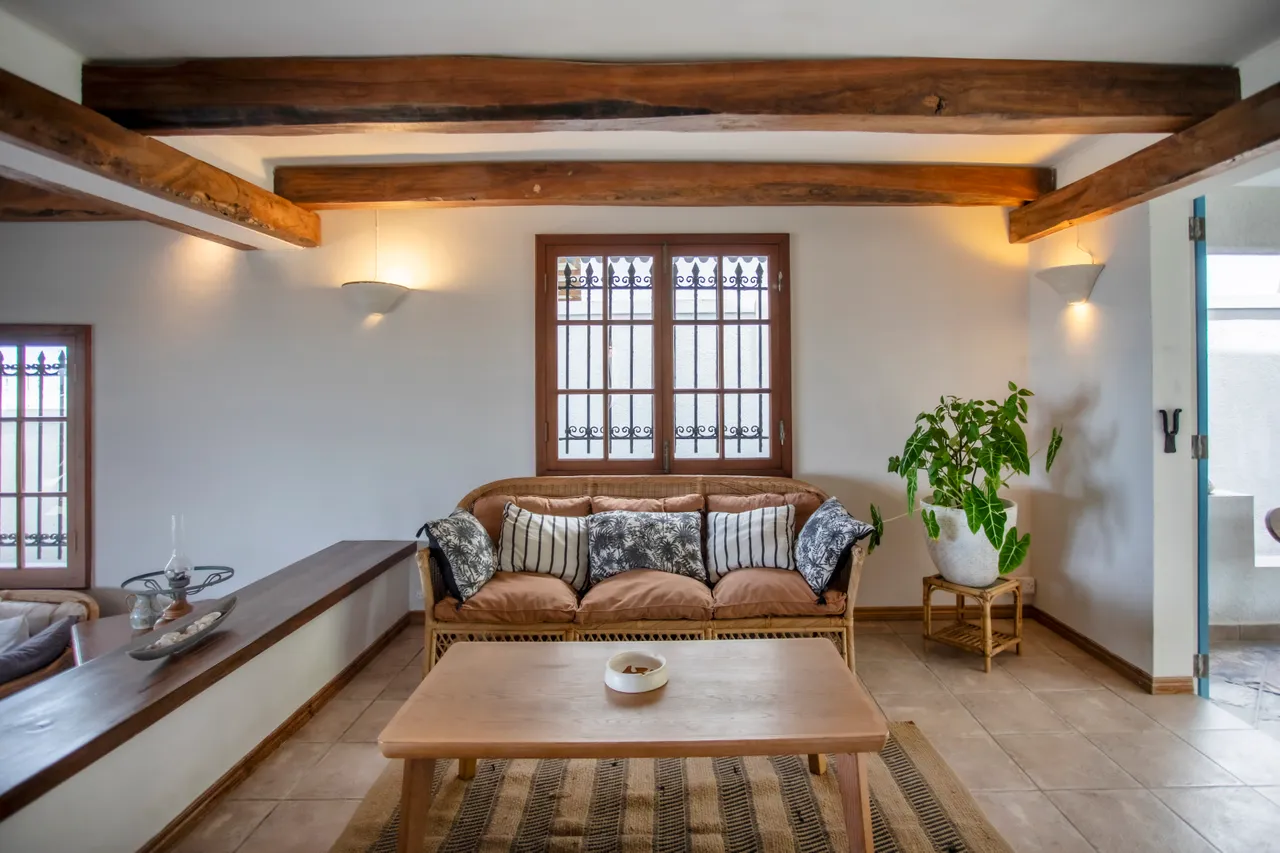 Galerie. Espace de vie lumineux avec canapé en rotin et table en bois. Plafond à poutres apparentes et grandes fenêtres.
