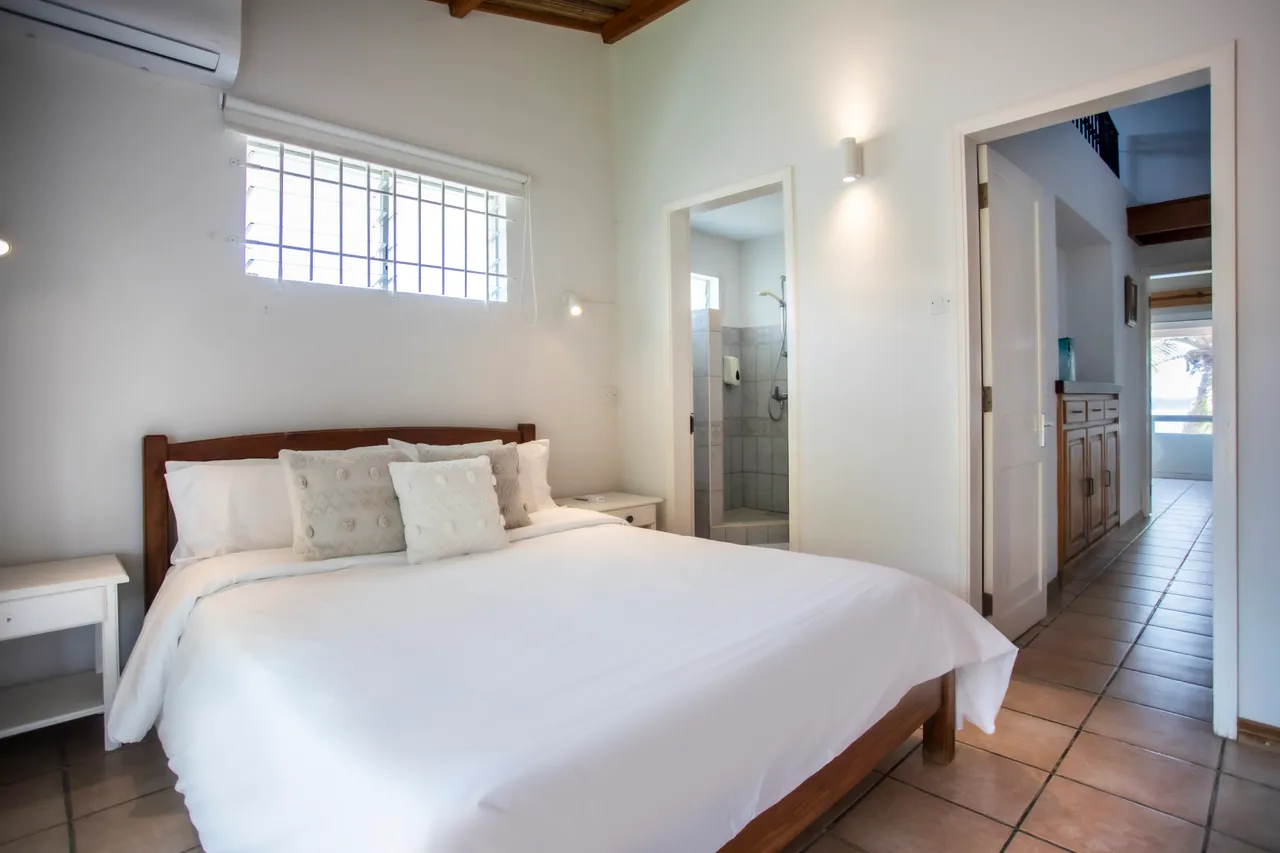 Chambre avec lit double et deux fenêtres, apportant une belle lumière naturelle. Décor simple avec mobilier en bois et carrelage au sol. Salle de bains attenante visible, offrant un accès pratique.