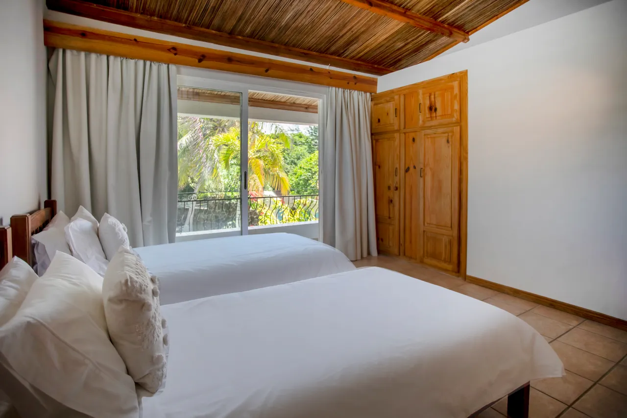 Teintes neutres avec touches de bois, la chambre dispose de deux lits simples. Les fenêtres offrent une vue sur la végétation. Placard en bois intégré pour le rangement. Plafond en bois et carrelage au sol.