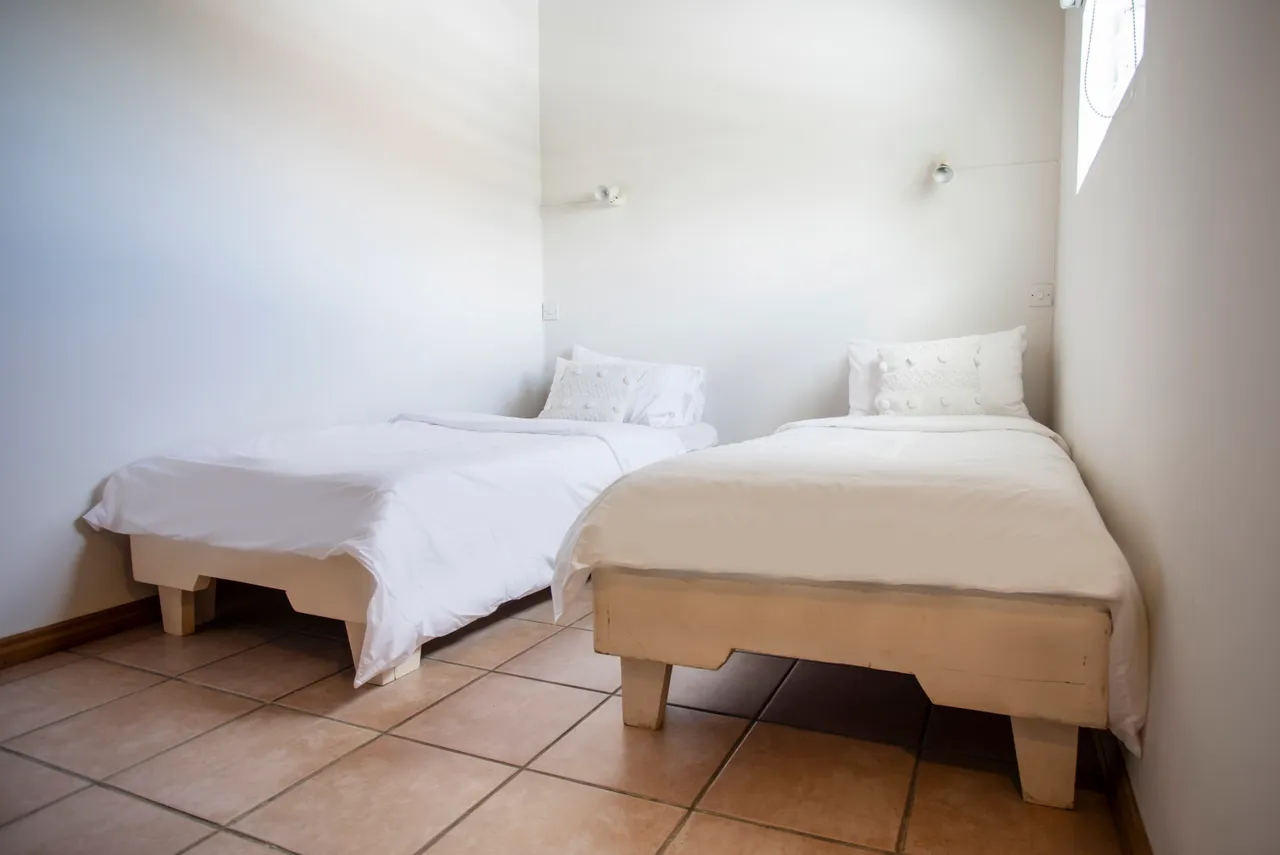 Chambre équipée de deux lits simples avec draps blancs. Éclairage naturel entrant par une fenêtre. Sol en carrelage clair et murs neutres. Rangements intégrés en bois disponibles.