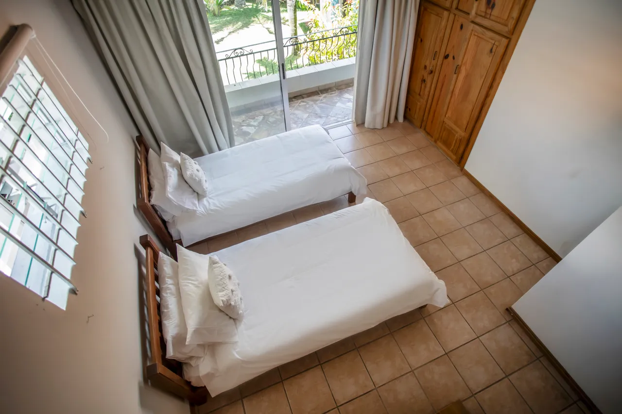 Teintes claires et sol en carrelage, la chambre dispose de deux lits simples. Fenêtre offrant une vue sur l'extérieur et rangements en bois intégrés. Plafond en bois accentuant l'espace.