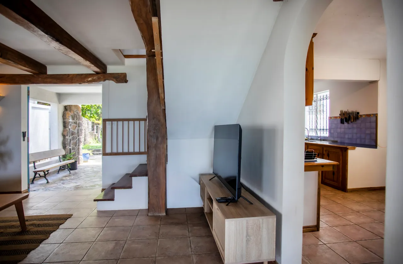 Galerie avec escalier en bois accentuant la hauteur sous plafond. Mobilier sobre et télévision. Sol en carrelage clair et murs peints.