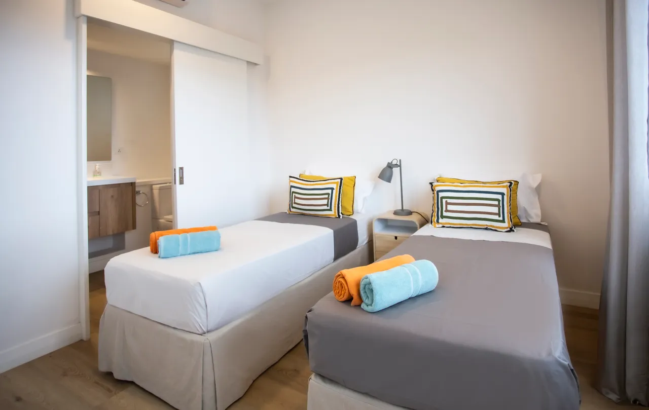 Chambre avec lits simples et salle de bain attenante.  
Mobilier en bois clair et décoration colorée pour une ambiance accueillante.  
Espace fonctionnel incluant un bureau et des rangements pratiques.  
Linge de lit et serviettes soigneusement disposés pour le confort.  
Lumière naturelle inondant la pièce grâce à une grande fenêtre.  
Ambiance ap