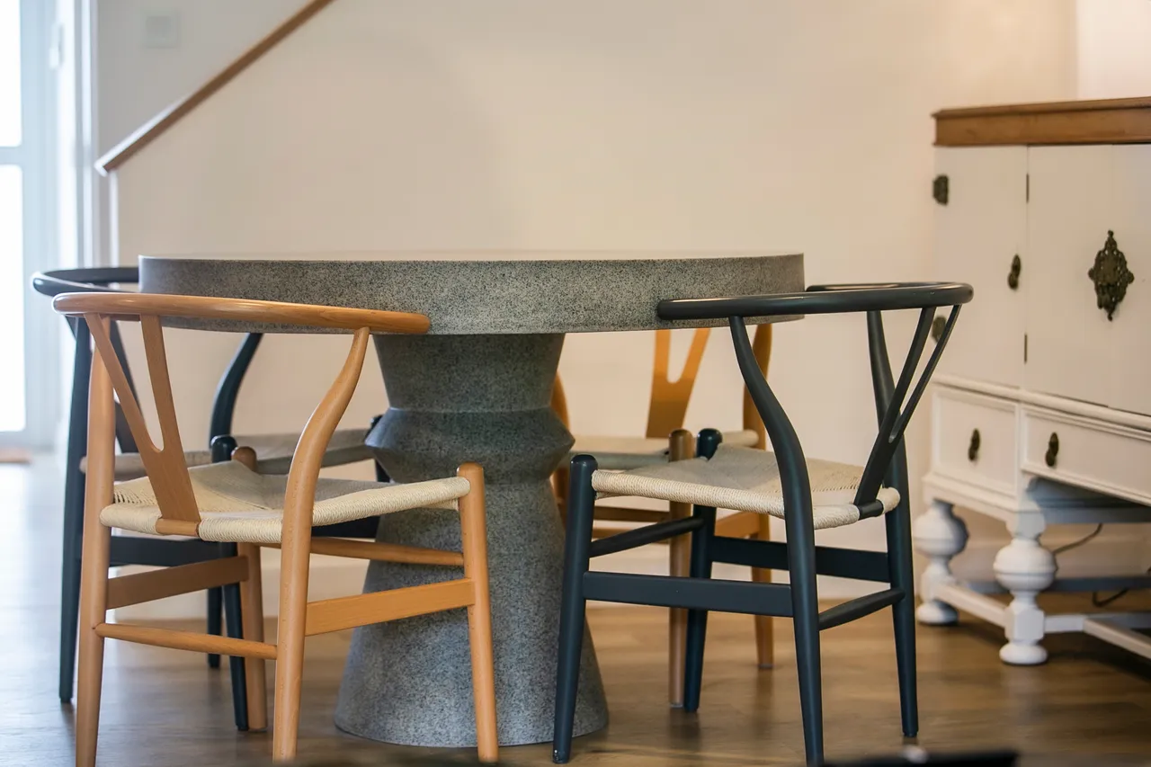 Table ronde avec piètement en pierre et chaises en bois.  
Textiles en corde tressée sur les assises ajoutent du confort.  
Éléments décoratifs en bois clair pour une ambiance naturelle.  
Sol en bois apportant chaleur et authenticité à l'espace.  
Lumière naturelle illuminant la pièce grâce aux grandes fenêtres.
