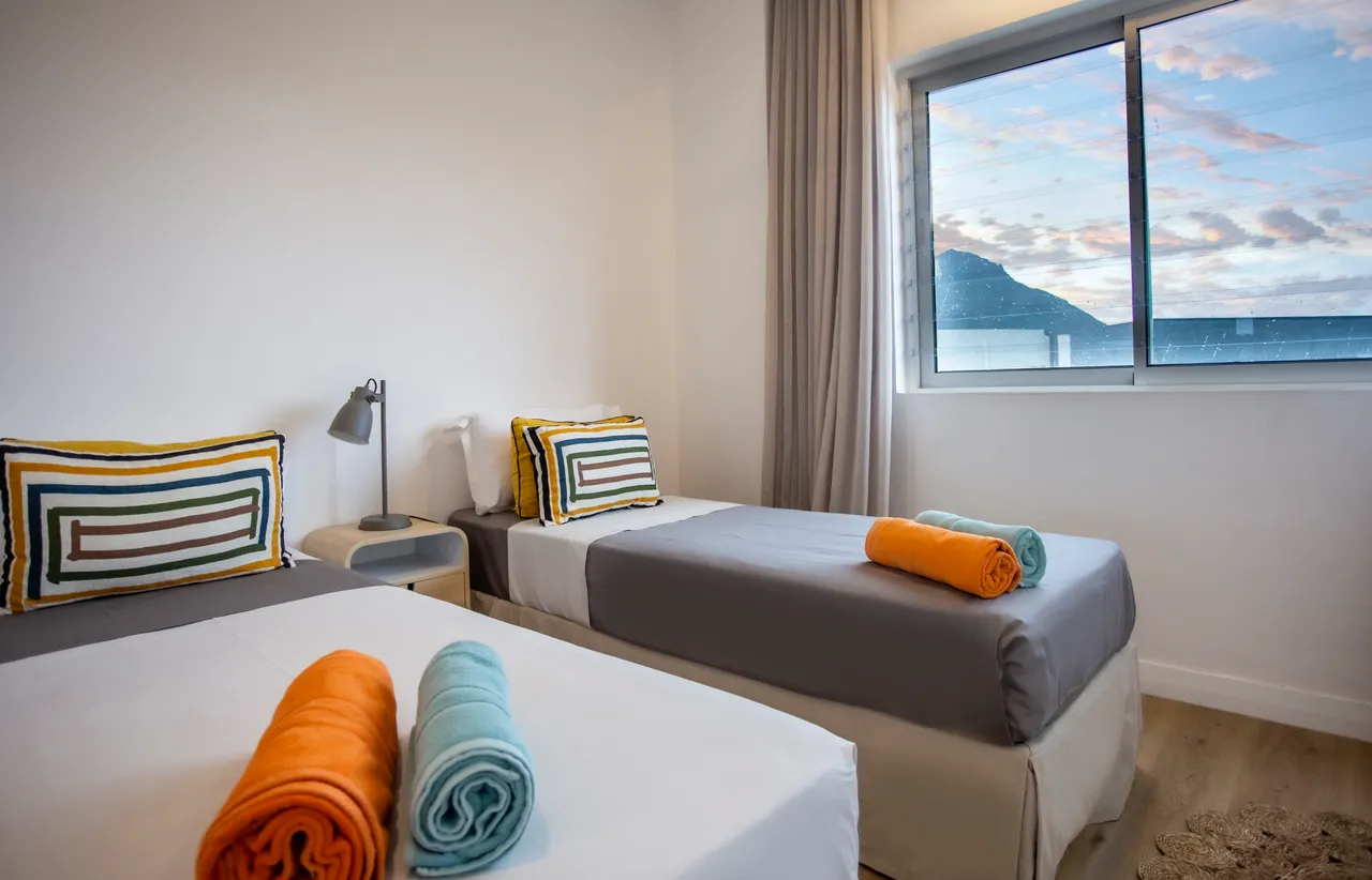 Chambre lumineuse avec deux lits simples et rangement pratique.  
Fenêtre offrant une vue dégagée sur l'extérieur.  
Coussins colorés ajoutent une touche de décoration.  
Éclairage moderne complétant l'espace fonctionnel.  
Linge de lit soigneusement disposé pour un aspect soigné.