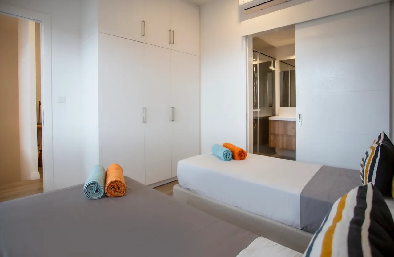 Chambre lumineuse avec deux lits simples et salle de bain attenante.  
Rangements intégrés et décoration colorée apportant du dynamisme à l'espace.  
Linge de lit disposé avec attention, créant une atmosphère soignée.  
Fenêtre laissant passer la lumière naturelle et offrant une vue sur l'extérieur.  
Ambiance apaisante grâce aux teintes douces du