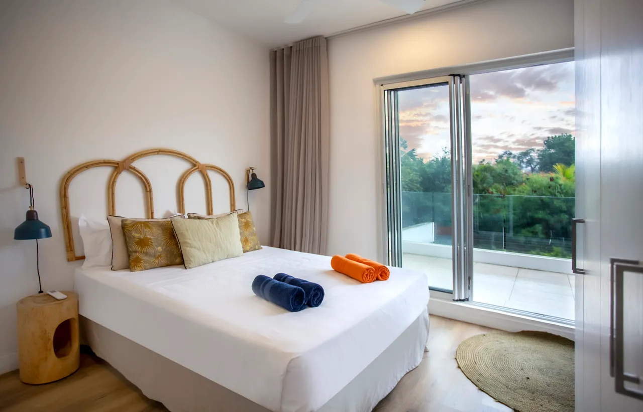 Chambre avec un lit double et accès direct à la terrasse.  
Éclairage doux provenant des lampes murales encadrant le lit.  
Serviettes colorées soigneusement disposées sur la literie.  
Fenêtre panoramique offrant une vue sur l'extérieur.  
Sol en bois créant une ambiance chaleureuse dans l'espace.