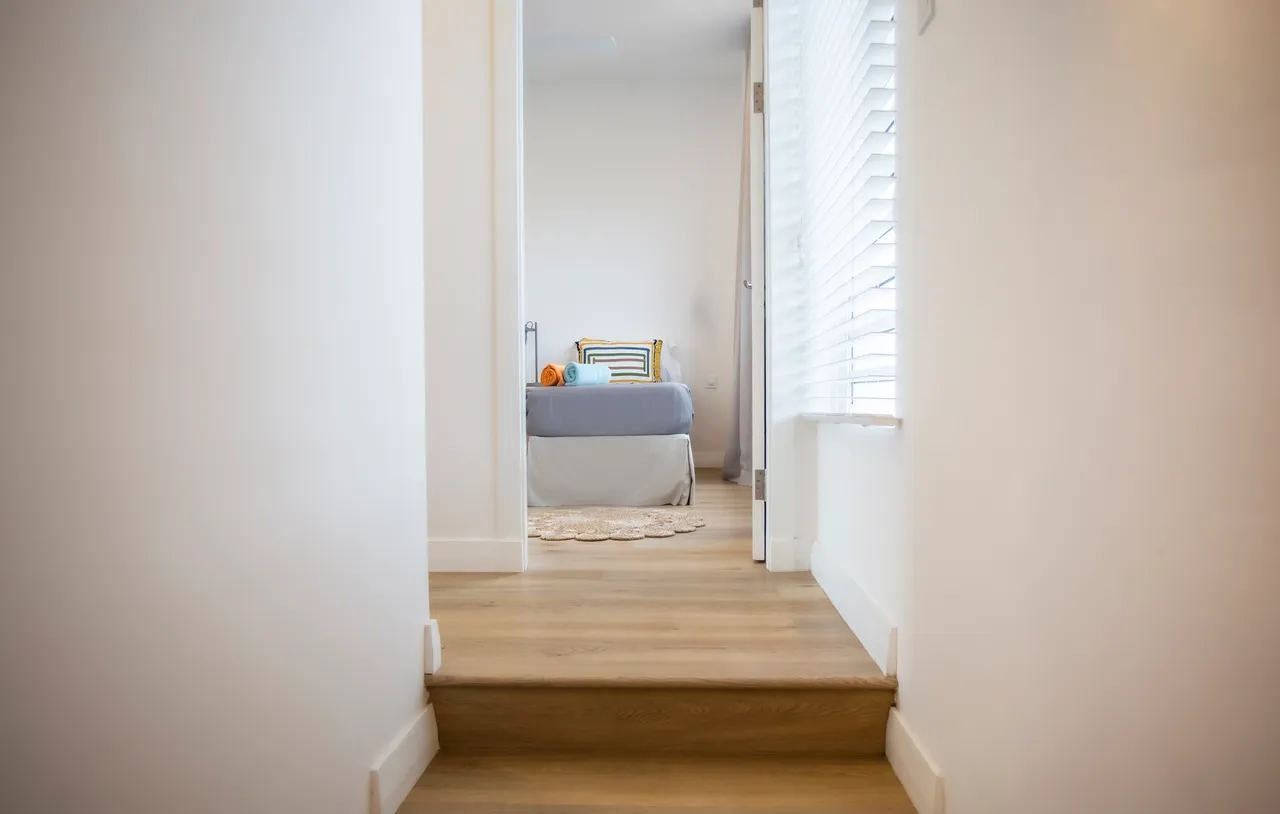 Tons clairs de beige et de blanc dans un couloir lumineux.  
Escalier en bois menant aux espaces de vie supérieurs.  
Fenêtre à lamelles apportant lumière naturelle et ventilation.  
Sol en bois harmonisant avec l’ambiance moderne du logement.  
Ambiance aérée grâce à un agencement minimaliste et épuré.