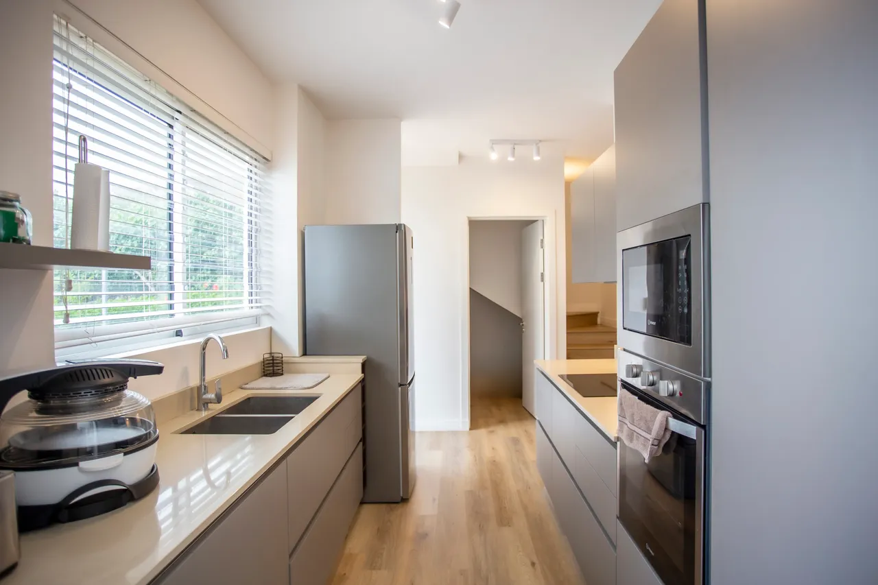 Cuisine moderne avec plans de travail clairs et rangement intégré.  
Fenêtre apportant lumière naturelle dans l'espace de préparation des repas.  
Appareils électroménagers récents facilitant la cuisson et le rangement.  
Éclairage fonctionnel au plafond pour une meilleure visibilité.  
Agencement optimisé pour une circulation fluide dans la cuisine.