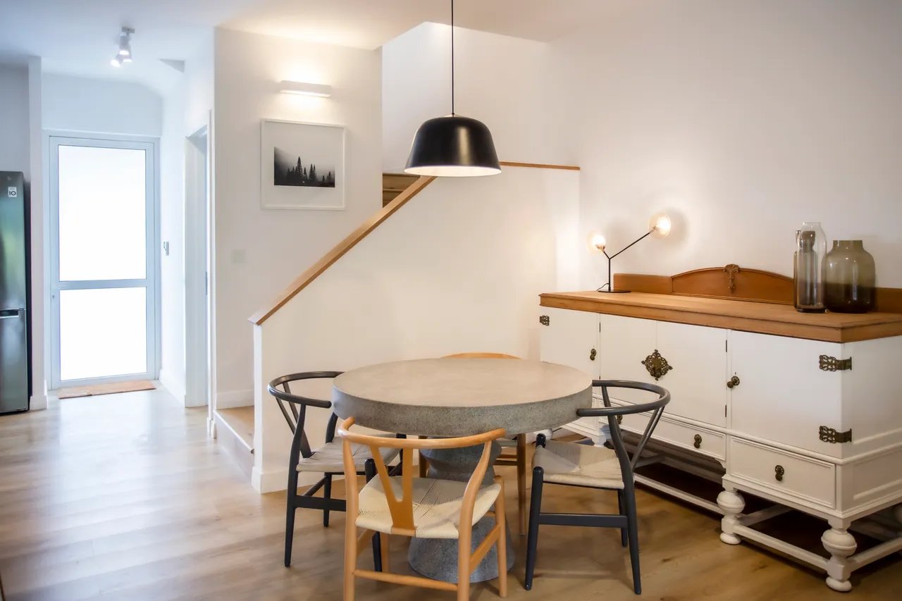 Espace de salle à manger avec une table ronde en pierre.  
Buffet en bois offrant du rangement pour la vaisselle.  
Éclairage suspendu noir ajoutant une note moderne à l'espace.  
Sol en bois clair créant une ambiance chaleureuse et contemporaine.  
Fenêtre laissant entrer la lumière naturelle et apportant une vue sur l'extérieur.