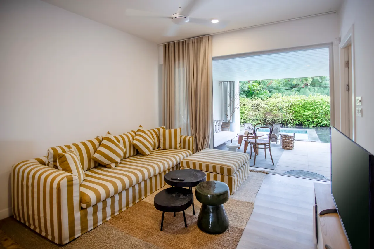 Espace de vie avec canapé à rayures et accès direct à la terrasse.  
Ventilateur de plafond offrant un confort supplémentaire dans la pièce.  
Table basse en bois ajoutant une fonctionnalité à l'aménagement.  
Rideaux clairs permettant à la lumière naturelle de filtrer.  
Vues sur le jardin depuis les fenêtres du salon.