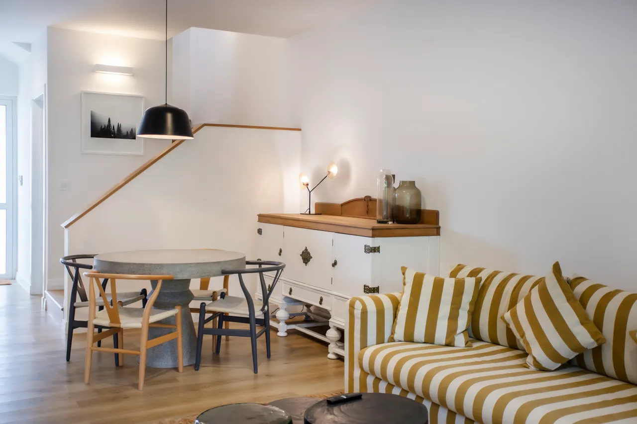 Espace de vie avec un canapé à rayures et une table en pierre.  
Buffet en bois offrant un espace de rangement pratique.  
Éclairage suspendu noir ajoutant une touche contemporaine.  
Sol en bois clair créant une atmosphère chaleureuse et aérée.  
Escalier menant aux niveaux supérieurs, intégrant le design de l'espace.