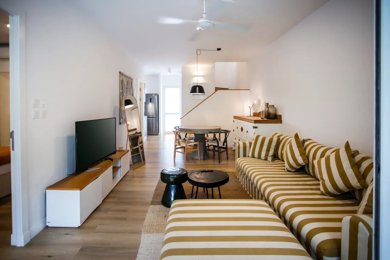 Espace de vie lumineux avec canapé à rayures et télévision.  
Ventilateur de plafond offrant une brise agréable dans l'espace.  
Table ronde en bois entourée de chaises, favorisant les repas conviviaux.  
Buffet blanc proposant un rangement pratique pour divers objets.  
Sol en bois clair renforçant la chaleur et l'ouverture de la pièce.