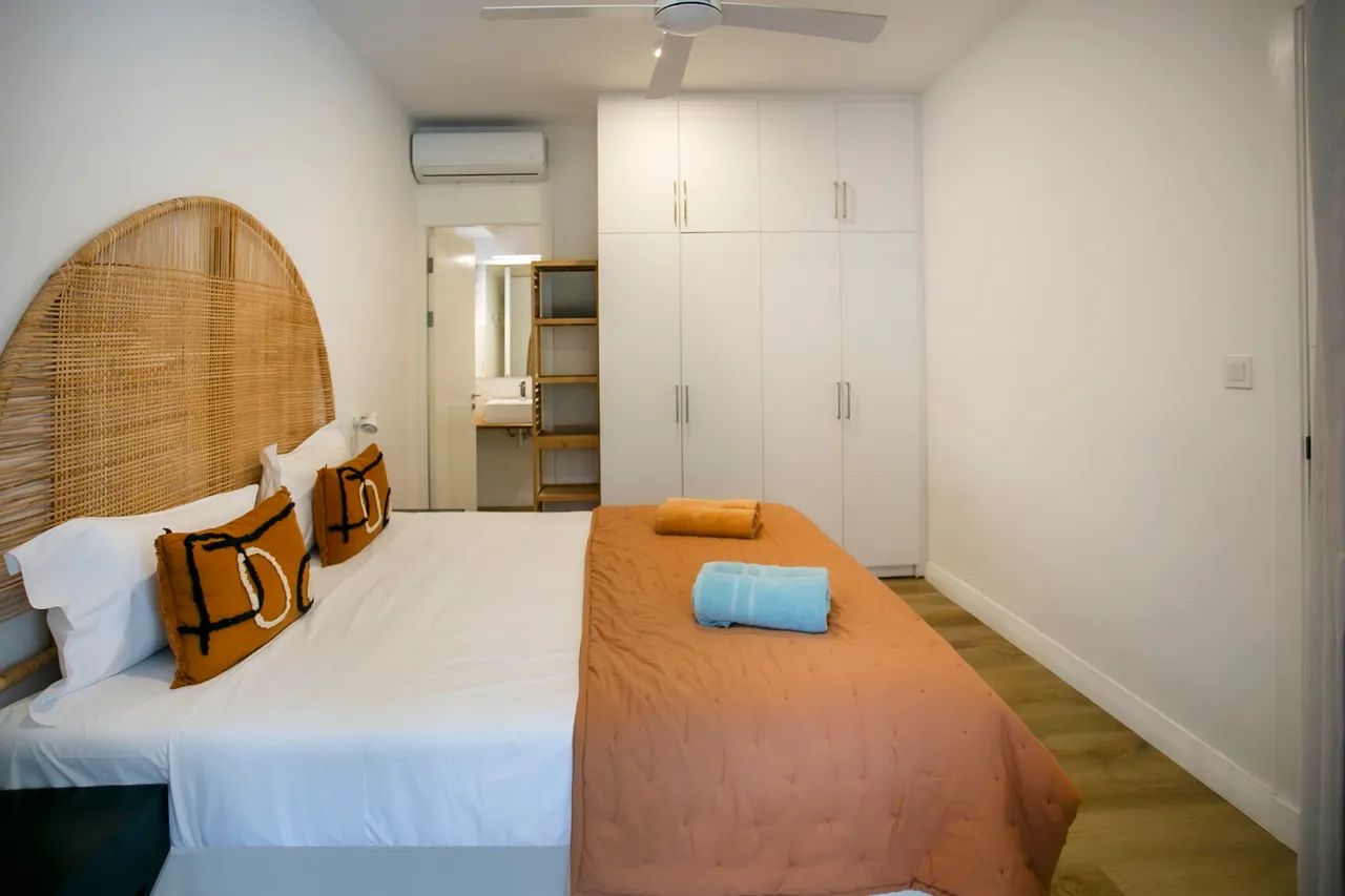 Chambre avec lit double et tête de lit en rotin.  
Éclairage discret près du lit pour une ambiance douce.  
Rangements intégrés avec meuble TV pratique et fonctionnel.  
Ventilateur de plafond assurant une circulation d'air agréable.  
Linge de lit coloré ajoutant une touche de contraste.