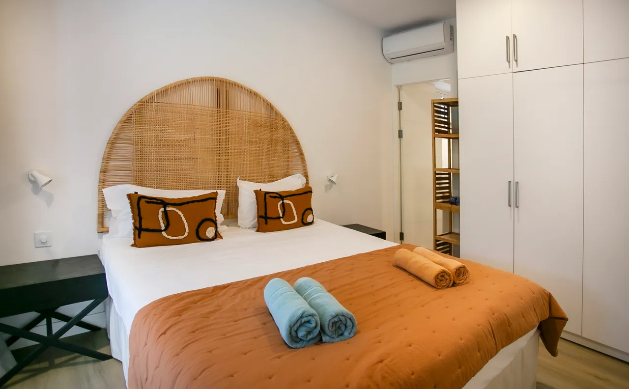 Chambre avec lit double et tête de lit en rotin.  
Éclairage doux sur les murs créant une atmosphère relaxante.  
Rangements intégrés offrant un espace fonctionnel et organisé.  
Linge de lit coloré ajoutant une touche de chaleur à la pièce.  
Climatisation assurant un confort thermique dans l'espace.
