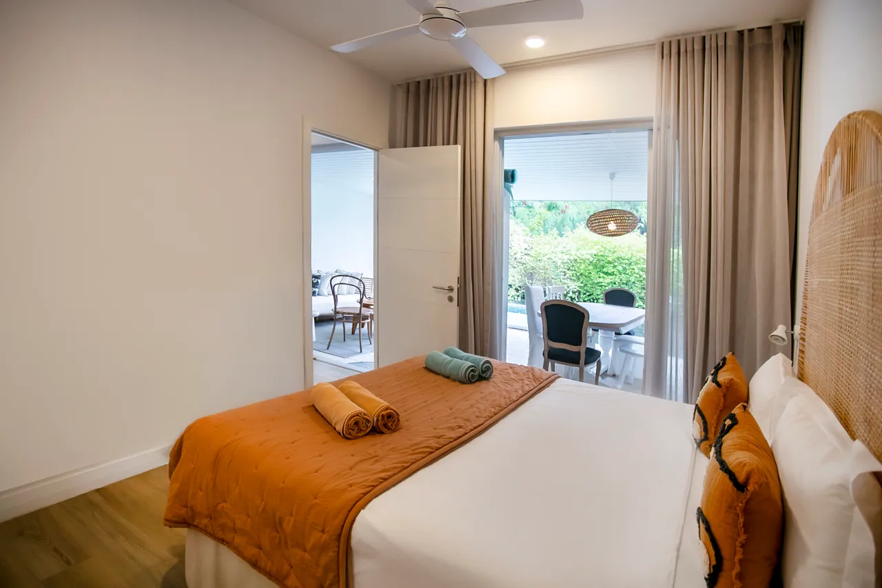 Chambre avec lit double et linge de lit aux couleurs chaleureuses.  
Accès direct à l'extérieur par des portes-fenêtres.  
Éclairage doux sur les murs créant une ambiance tranquille.  
Ventilateur de plafond assurant une circulation d'air agréable.  
Rangements intégrés pour une organisation optimale des effets personnels.