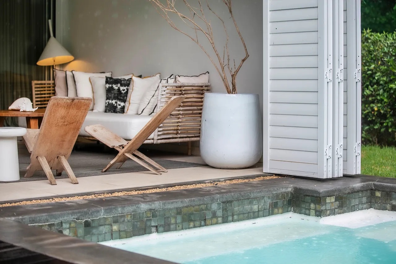 Espace extérieur avec mobilier en bois, favorisant la convivialité.  
Vue directe sur la piscine, ajoutant une dimension rafraîchissante.  
Ambiance calme grâce à la végétation environnante.  
Sol carrelé pour un entretien simple et efficace.  
Éléments décoratifs minimalistes créent une atmosphère apaisante.