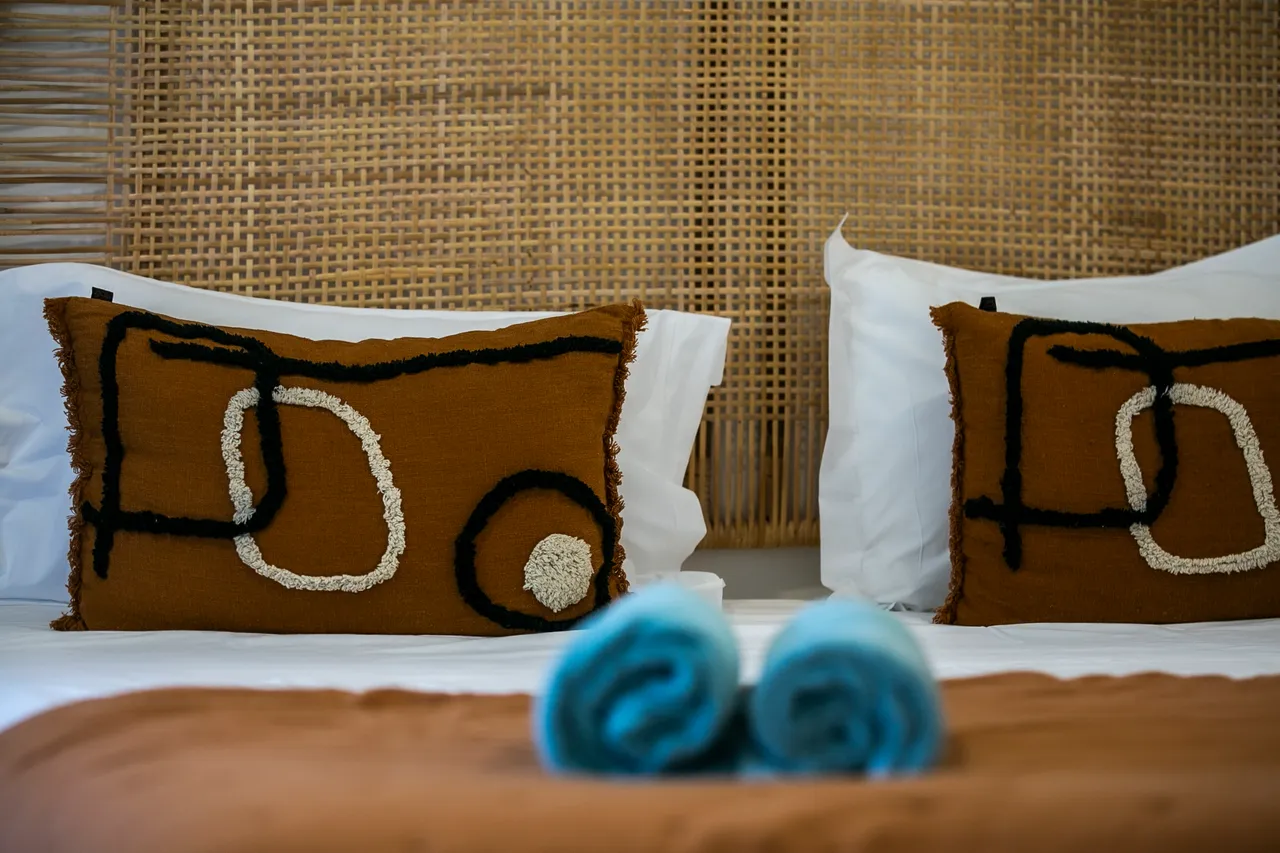 Textiles aux motifs organiques sur les coussins du lit.  
Tôles en bambou ajoutent une texture naturelle et un aspect chaleureux.  
Couleurs douces créant une ambiance sereine dans la chambre.  
Serviettes roulées sur la literie, offrant une touche de confort.  
Linge de lit clair pour une esthétique épurée et reposante.