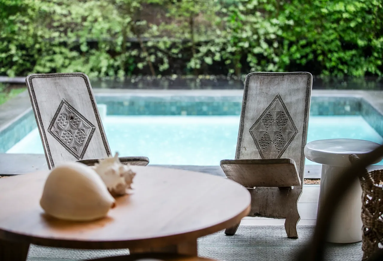Chaises en bois avec motifs sculptés, offrant un coin de repos.  
Vue sur la piscine à proximité, enrichissant l'espace extérieur.  
Table ronde en bois ajoutant une touche naturelle à l'agencement.  
Éléments de décoration simples créant une ambiance apaisante.  
Végétation luxuriante entourant l'espace pour plus d'intimité.