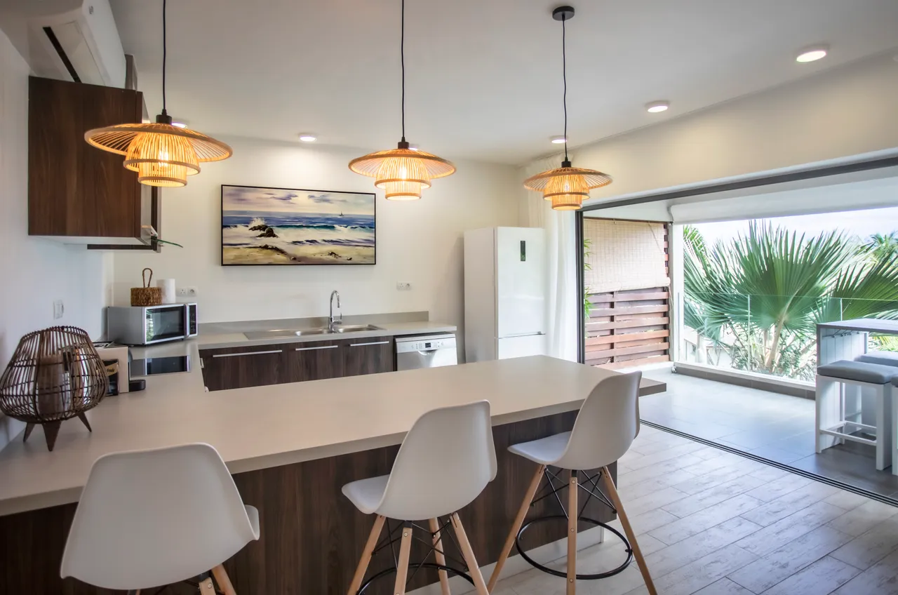 Commodités modernes avec bar à petit-déjeuner et chaises contemporaines.  
Éclairage suspendu en osier ajoutant une touche chaleureuse à l'espace.  
Cuisine ouverte sur le salon, favorisant les interactions.  
Fenêtres donnant sur l'extérieur, apportant lumière naturelle et vue.  
Plan de travail spacieux offrant un espace fonctionnel pour cuisiner.