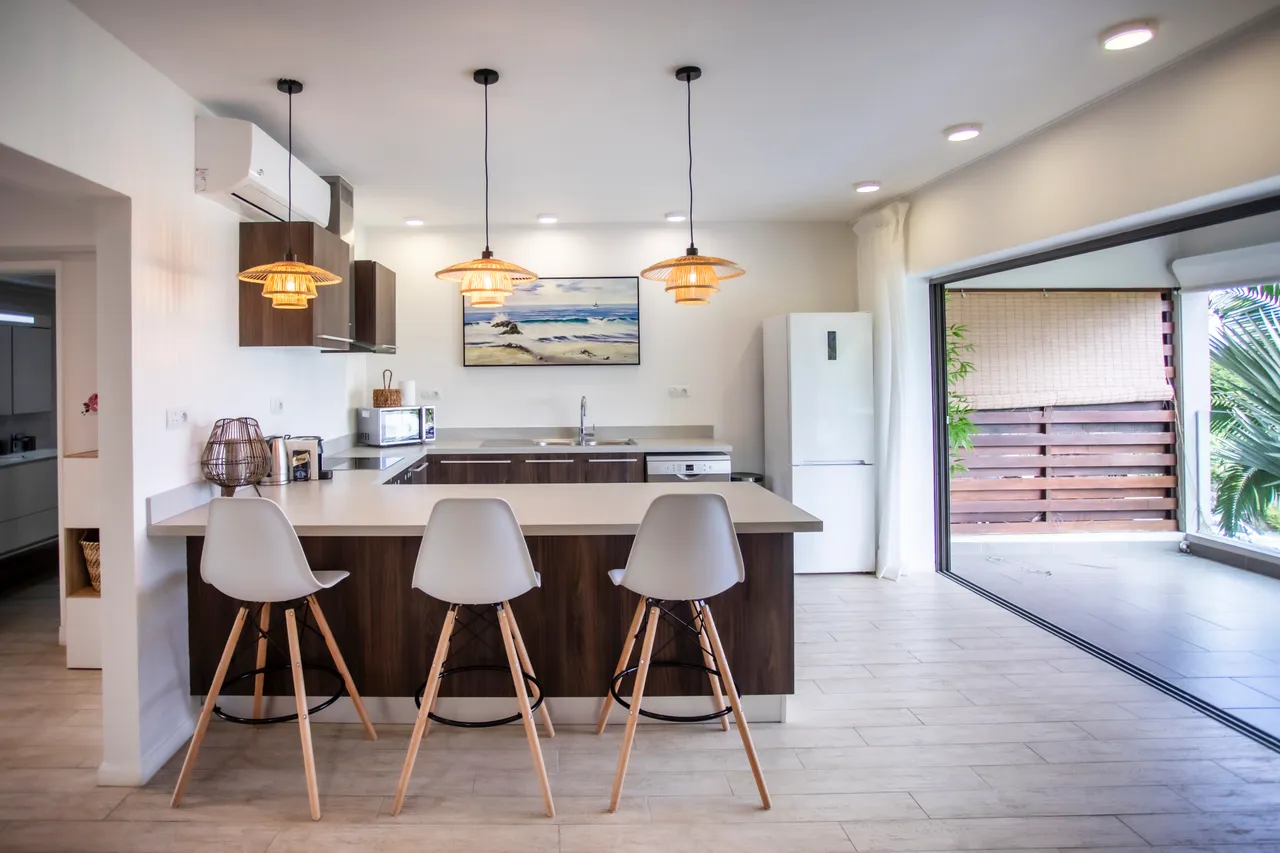 Cuisine moderne avec un bar à petit-déjeuner et éclairage suspendu.  
Espace fonctionnel avec plan de travail spacieux pour préparer des repas.  
Fenêtres laissant entrer la lumière naturelle et offrant une vue sur l'extérieur.  
Mobilier en bois et chaises contemporaines créant une ambiance accueillante.  
Accès direct à l'extérieur depuis la cuisine, facilitant la circulation.