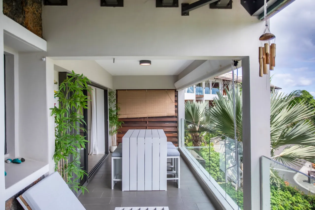 Terrasse couverte avec mobilier blanc et vue sur le jardin.  
Végétation verte ajoutant une touche de fraîcheur à l'espace.  
Éclairage naturel grâce aux grandes fenêtres offrant une vue dégagée.  
Mobilier moderne en bois créant une atmosphère conviviale sur la terrasse.  
Accès direct depuis les espaces intérieurs, facilitant la circulation.