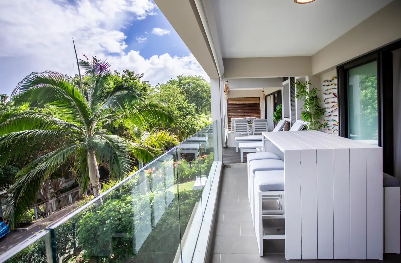 Terrasse spacieuse avec mobilier contemporain offrant une vue sur le jardin.  
Végétation luxuriante visible depuis le balcon, créant une ambiance naturelle.  
Espace extérieur lumineux grâce aux grandes fenêtres permettant une bonne luminosité.  
Coin repas sur la terrasse, propice aux repas en plein air.  
Accès direct depuis le salon, facilitant la circulation entre les espaces.