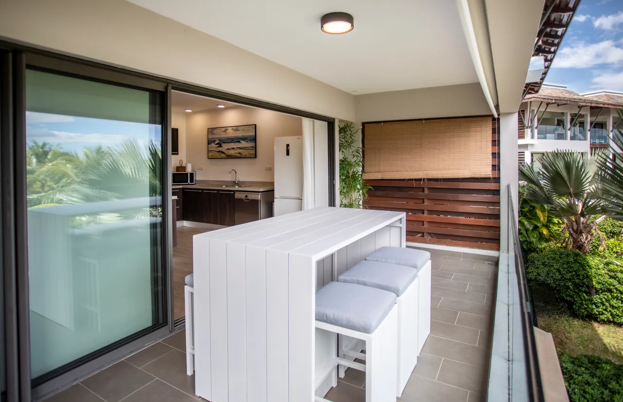 Surface claire avec un bar extérieur et chaises modernes.  
Vue sur le jardin verdoyant depuis l'espace de repas.  
Accès direct à la cuisine favorisant des échanges conviviaux.  
Matériaux contemporains ajoutant une touche de style à l'extérieur.  
Ambiance détendue créant un lieu agréable pour se réunir.