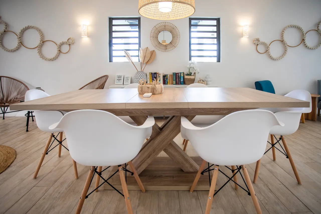Table à manger en bois avec chaises modernes blanches, créant un espace convivial.  
Éclairage suspendu en osier apportant une ambiance chaleureuse à la pièce.  
Éléments décoratifs naturels ajoutant une touche organique à l'ensemble.  
Fenêtres latérales offrant une luminosité naturelle dans l'espace.  
Agencement ouvert facilitant les interactions entre les différents espaces.