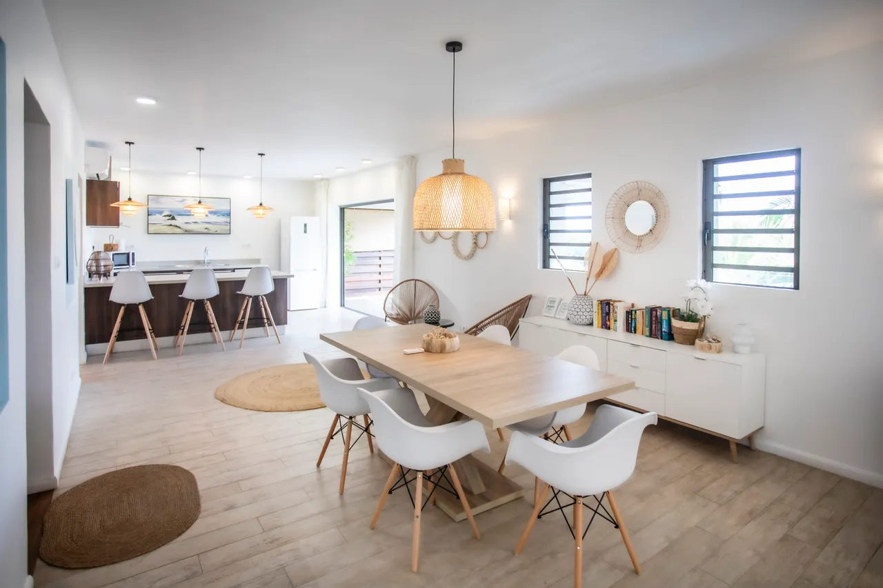Tons clairs et chaleureux dans l'espace de vie avec des éléments en bois.  
Salle à manger spacieuse avec une grande table et chaises modernes.  
Éclairage suspendu en osier ajoutant une touche naturelle à la pièce.  
Fenêtres latérales apportant luminosité et ventilation à l'intérieur.  
Agencement ouvert reliant le coin repas et la cuisine de manière fluide