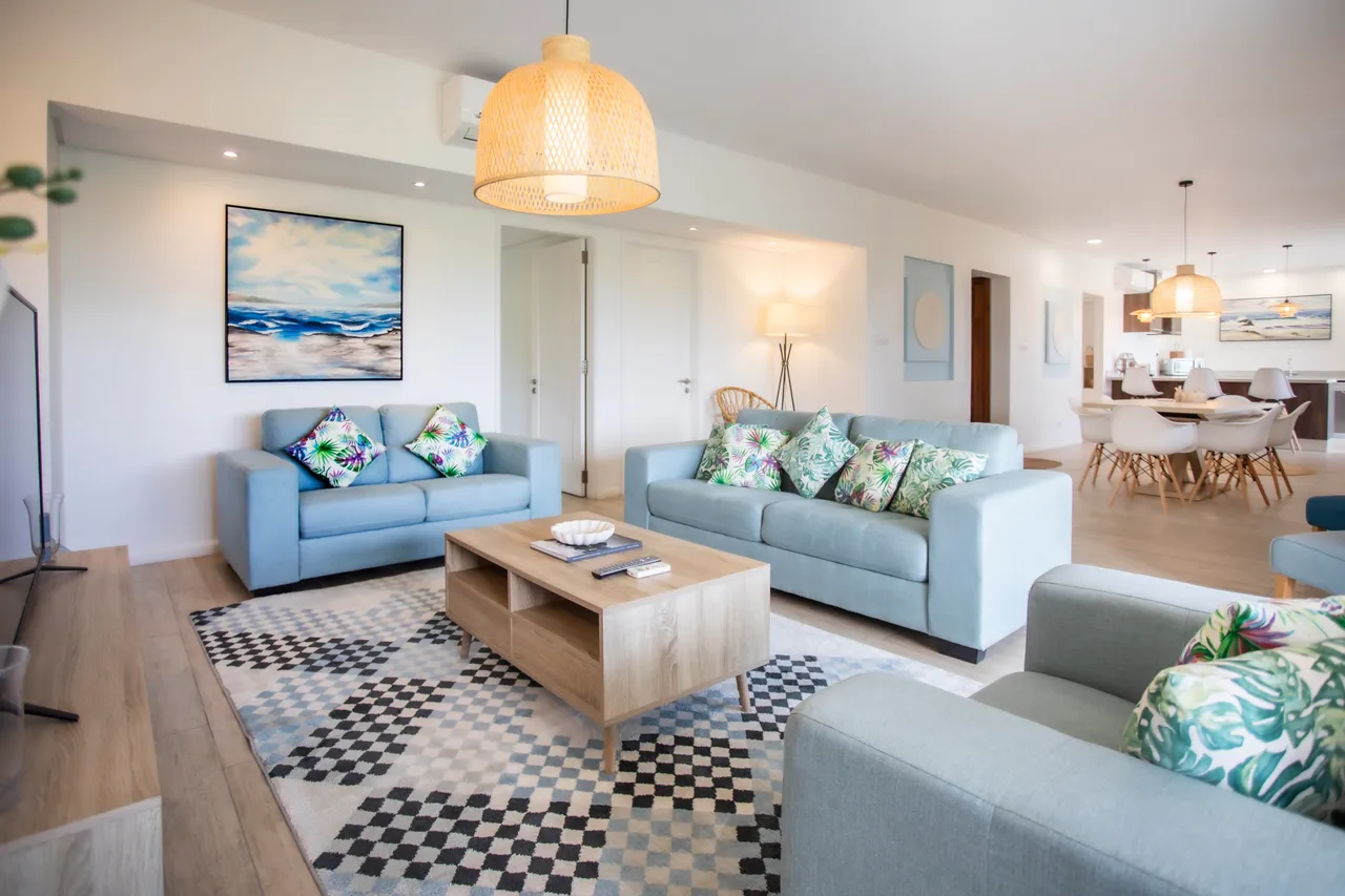 Espace de vie avec canapés clairs et tapis graphique au sol.  
Éclairage suspendu en osier ajoutant une ambiance chaleureuse.  
Mobilier moderne créant un cadre contemporain pour la détente.  
Grandes fenêtres offrant une bonne luminosité et vue sur l'extérieur.  
Agencement ouvert reliant le salon à la salle à manger.