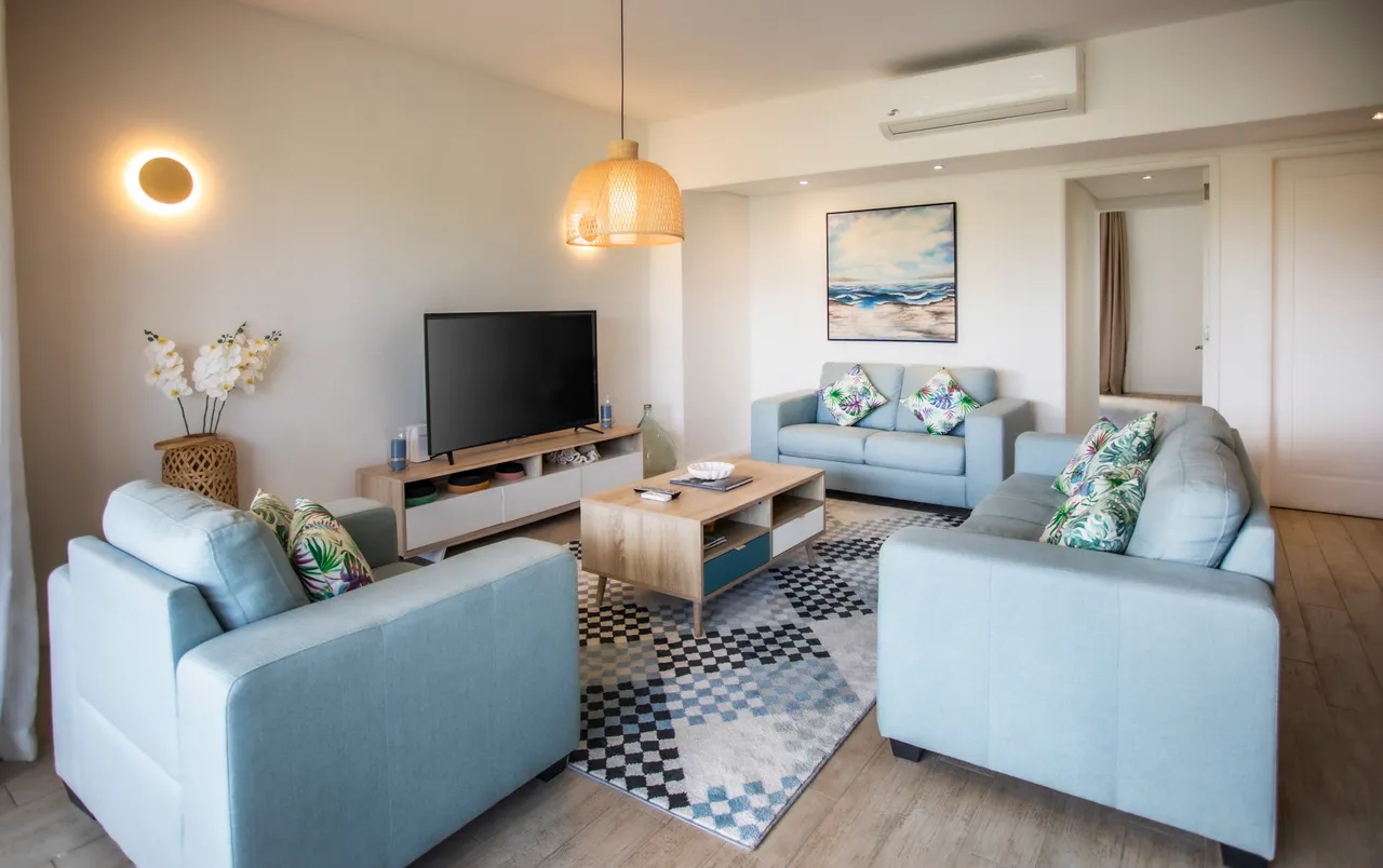 Espace de salon avec canapés clairs et table basse en bois.  
Éclairage suspendu en osier apportant une chaleur à l'intérieur.  
Grandes fenêtres assurant une bonne luminosité et vue sur l'extérieur.  
Décor minimaliste avec touches de couleur dans les coussins.  
Agencement ouvert facilitant l'accès aux différentes zones de l'appartement.