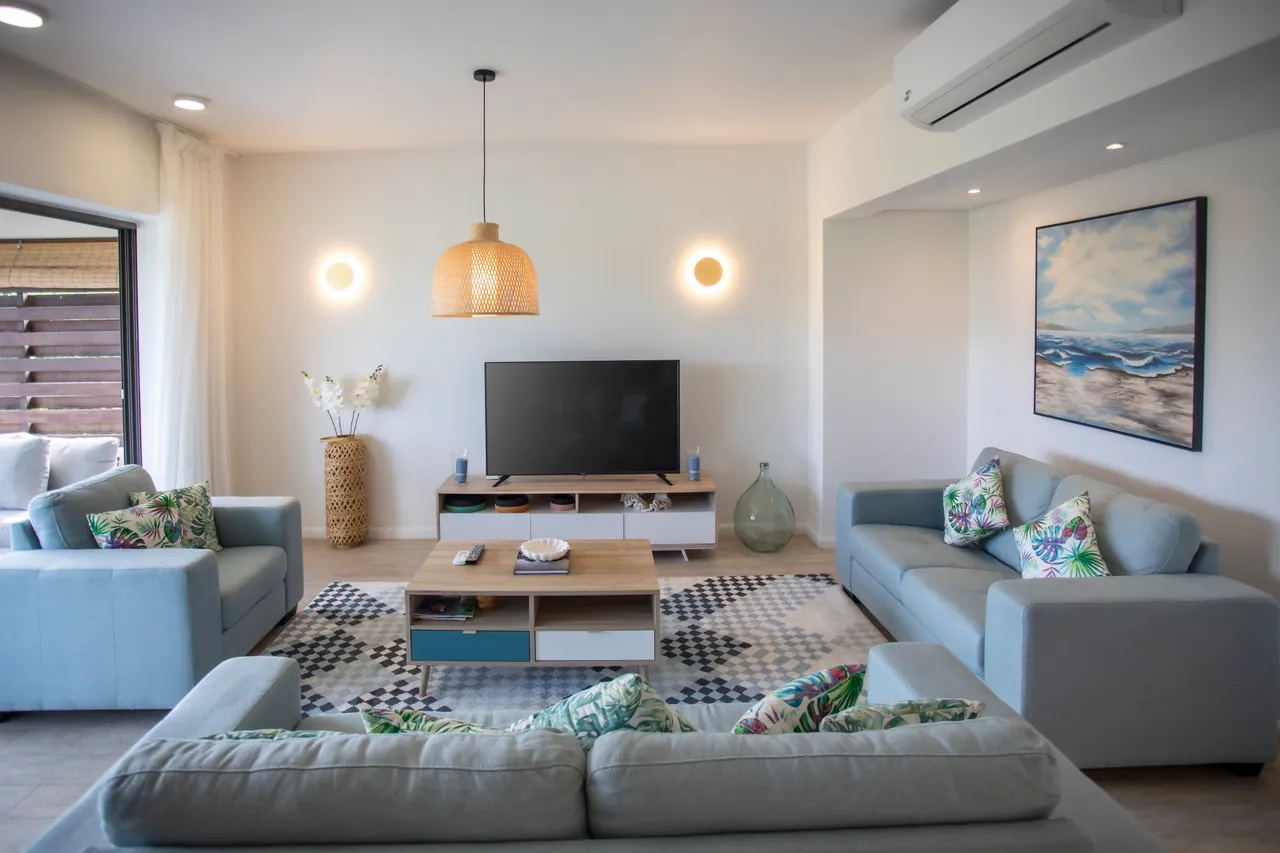 Tons clairs et frais des canapés contrastent avec le tapis graphique.  
Éclairage naturel abondant grâce aux grandes baies vitrées.  
Mobilier moderne en bois créant un espace de vie fonctionnel.  
Accessibilité directe à la terrasse depuis l'espace de salon.  
Coussins colorés ajoutent une touche de vie à l'ambiance intérieure.