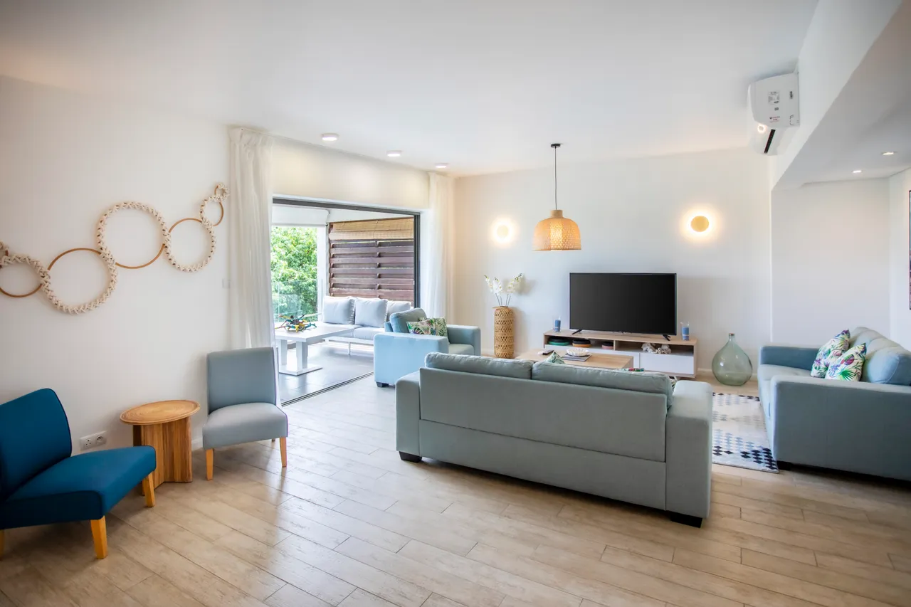 Espace de salon avec un accès direct à la terrasse extérieure.  
Éclairage suspendu en osier ajoutant une note chaleureuse à l'intérieur.  
Canapés clairs offrant un cadre confortable pour la détente.  
Grandes baies vitrées assurant une lumière naturelle abondante.  
Décor minimaliste avec des éléments en bois intégrés.