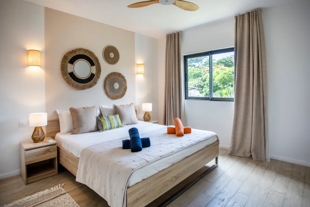 Chambre avec lit double et décoration murale en osier.  
Grandes fenêtres laissant entrer la lumière naturelle.  
Linge de lit aux teintes douces et coussins colorés.  
Mobilier en bois créant une ambiance chaleureuse et contemporaine.  
Éclairage mural apportant une touche douce à la pièce.