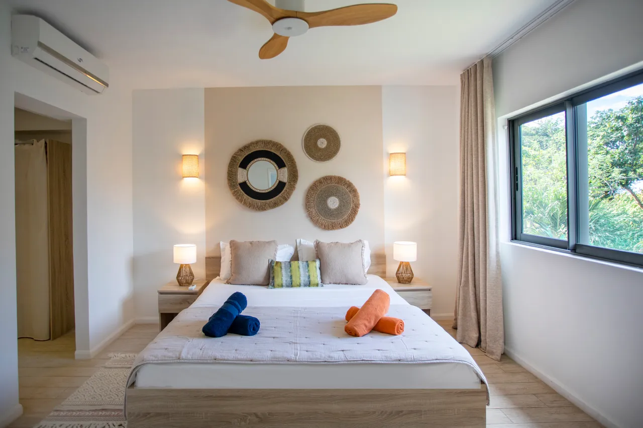 Chambre spacieuse avec de grandes fenêtres offrant une vue sur la nature.  
Lit double avec tête de lit en bois et coussins colorés.  
Éclairage doux apportant une ambiance apaisante à l'espace.  
Mobilier en bois intégrant des éléments décoratifs contemporains.  
Ventilateur au plafond pour un confort optimal pendant les nuits chaudes.