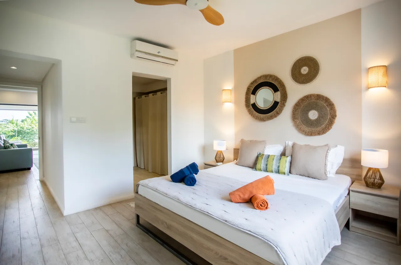 Chambre avec lit double et décor mural en osier naturel.  
Grandes fenêtres permettant une bonne luminosité et vue sur l'extérieur.  
Mobilier en bois ajoutant une touche contemporaine à l'espace.  
Éclairage doux créant une ambiance apaisante pour la détente.  
Ventilateur au plafond pour un confort agréable durant la nuit.