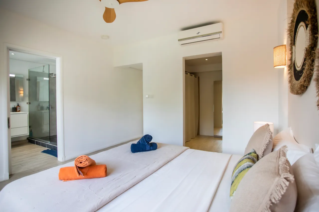 Chambre avec lits jumeaux et décoration murale en osier.  
Grandes fenêtres apportant une belle luminosité à l'espace.  
Linge de lit aux couleurs vives ajoutant une touche de vie.  
Mobilier en bois offrant un agencement fonctionnel et moderne.  
Éclairage mural créant une atmosphère douce et apaisante.