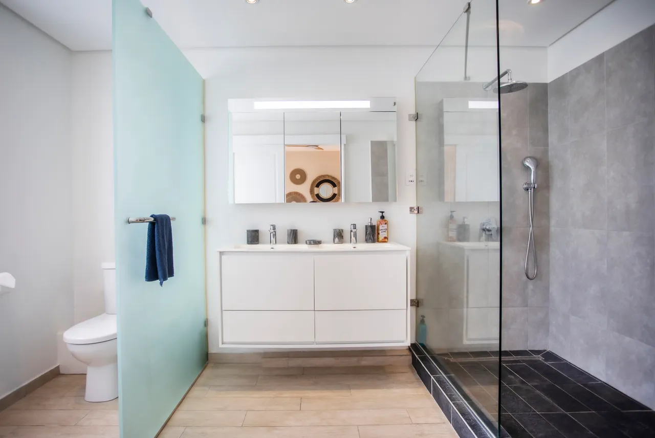 Salle de bain moderne avec douche spacieuse et parois en verre.  
Meuble lavabo blanc intégré offrant un espace de rangement.  
Éclairage LED au-dessus du miroir pour une bonne visibilité.  
Carrelage en grès cérame assurant une surface facile d'entretien.  
Tapis de bain en textile doux ajoutant un confort supplémentaire.