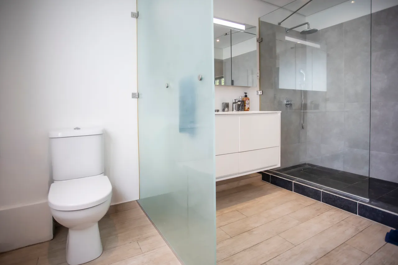 Salle de bain lumineuse avec douche à l'italienne et parois en verre.  
Meuble de rangement moderne avec un lavabo intégré pour plus de praticité.  
Éclairage LED au-dessus du miroir offrant une visibilité optimale.  
Carrelage clair et facile d'entretien pour un usage quotidien.  
Espace bien agencé pour un confort fonctionnel.