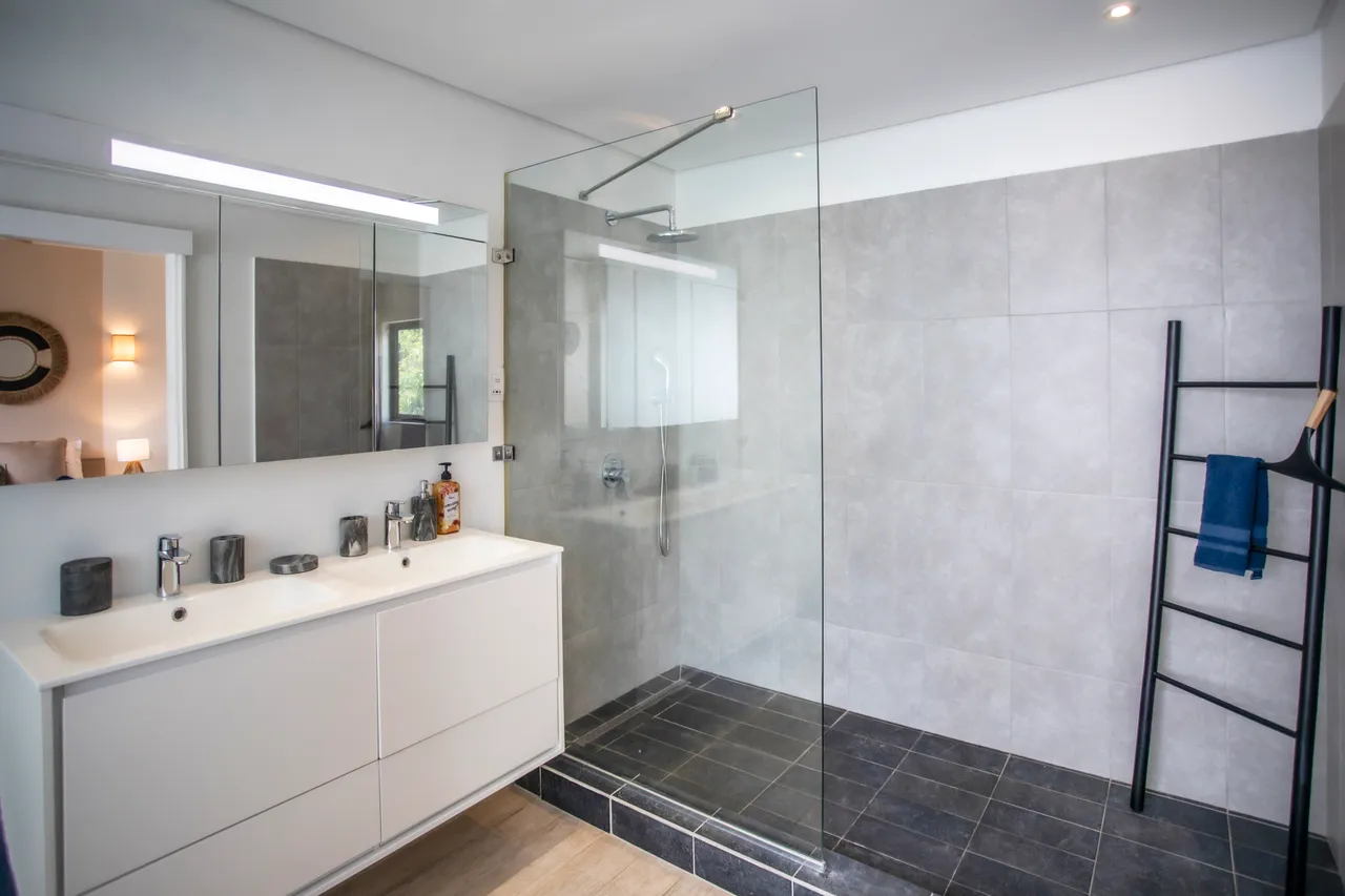 Salle de bain spacieuse avec douche italienne et parois en verre.  
Meuble de rangement pratique avec lavabo intégré pour un usage fonctionnel.  
Éclairage LED au-dessus du miroir garantissant une bonne visibilité.  
Carrelage en grès clair facilitant l'entretien quotidien.  
Détails soignés ajoutant une touche moderne à l'espace.