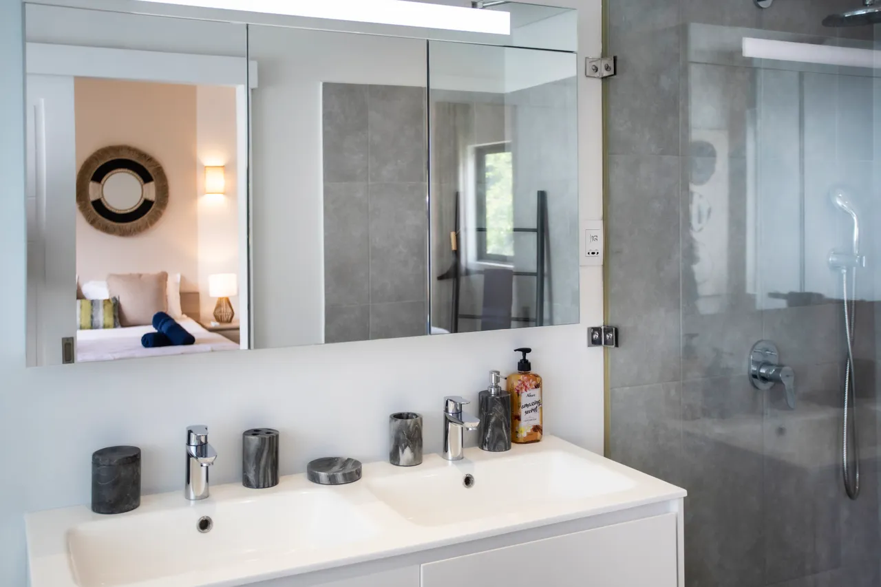 Salle de bain avec double vasque et grand miroir sur le mur.  
Douche à l'italienne avec parois en verre offrant une ambiance moderne.  
Éclairage LED au-dessus du miroir garantissant une visibilité optimale.  
Carrelage clair et élégant facilitant l'entretien quotidien.  
Accessoires en pierre ajoutant une touche de sophistication à l'espace.