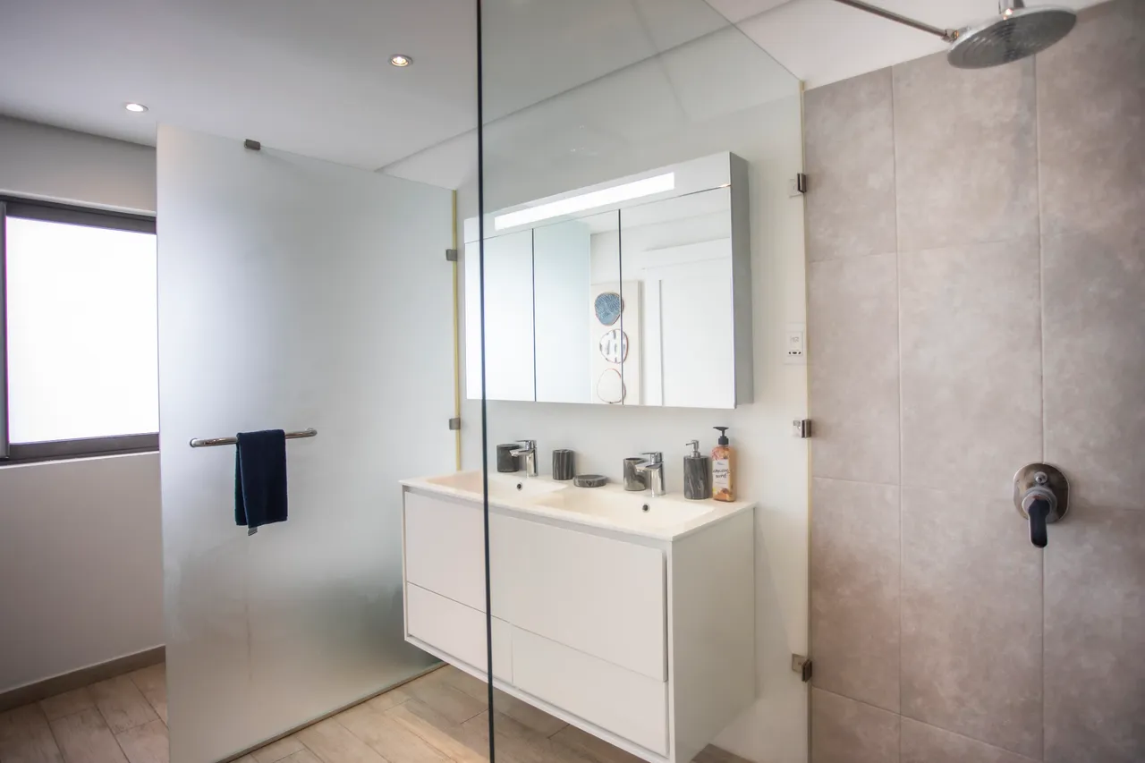 Salle de bain bien agencée avec double vasque et miroir éclairé.  
Douche à l'italienne avec parois en verre pour une ambiance moderne.  
Éclairage au-dessus du miroir assurant une visibilité optimale.  
Revêtement en carrelage clair facilitant l'entretien quotidien.  
Détails contemporains ajoutant une touche fonctionnelle à l'espace.