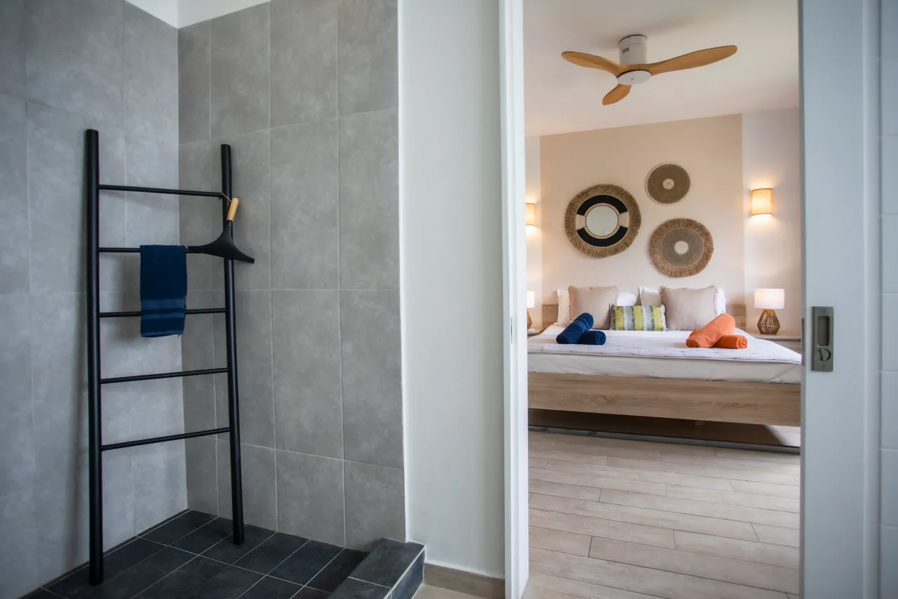 Salle de bain avec douche à l'italienne et carrelage clair.  
Échelle en métal pour suspendre les serviettes à proximité.  
Miroir arrondi et éclairage mural ajoutant une touche moderne.  
Ventilateur au plafond pour assurer une circulation d'air agréable.  
Accès direct à la chambre depuis l'espace de douche.