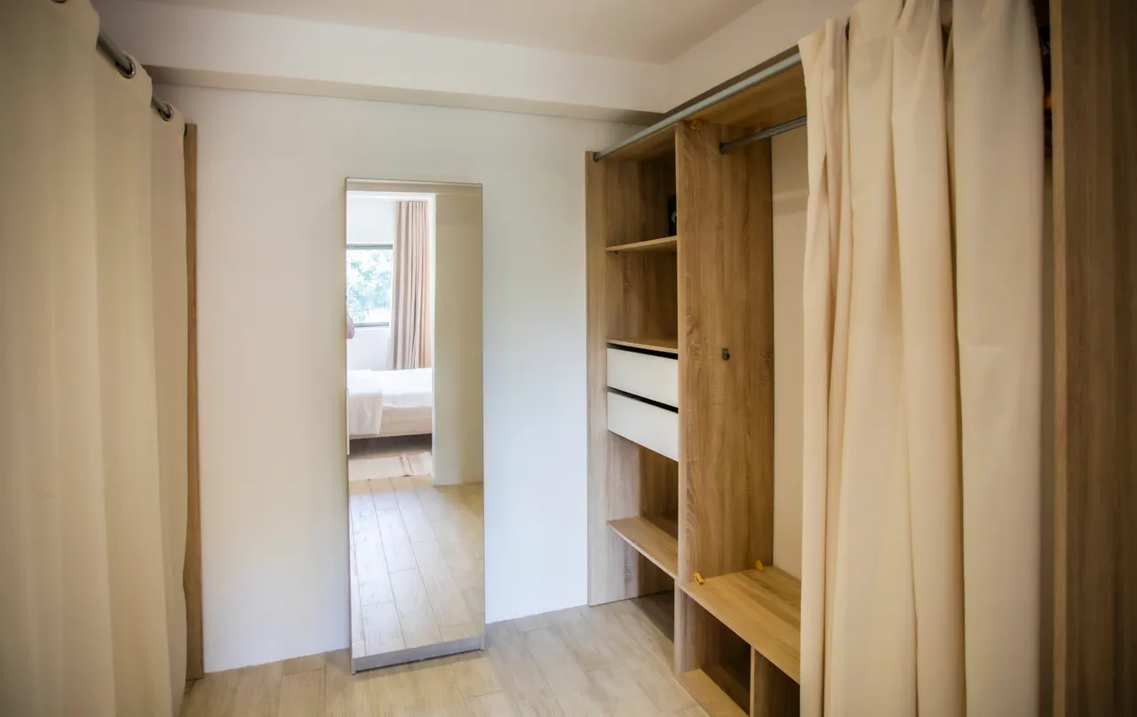 Espace de rangement avec étagères ouvertes et miroir intégré.  
Cloison en rideau séparant le dressing de la chambre.  
Lumière naturelle apportée par la fenêtre adjacente.  
Sol en bois offrant une atmosphère chaleureuse et contemporaine.  
Détails en bois naturel ajoutant une touche moderne au design.