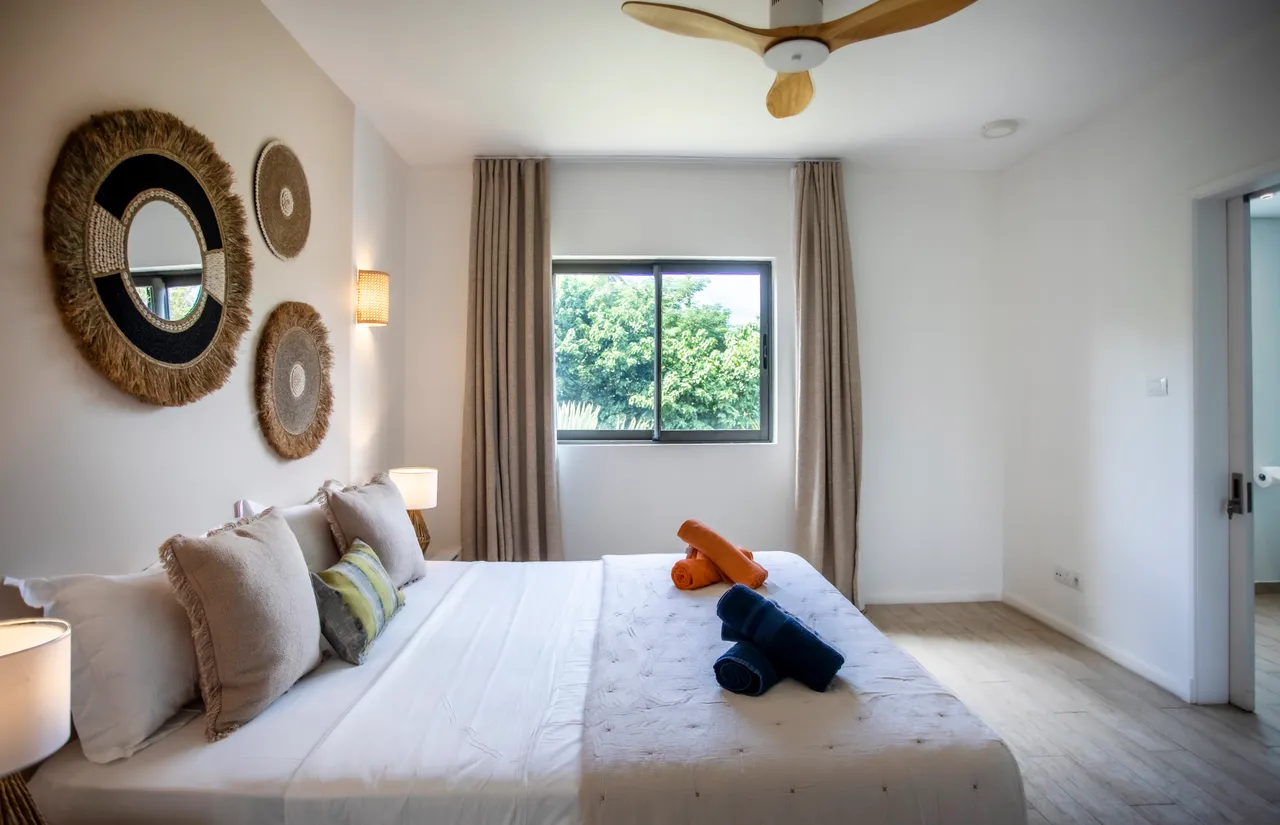 Chambre avec lit double agrémenté de coussins colorés.  
Éclairage mural et ventilateur de plafond pour une atmosphère agréable.  
Grande fenêtre offrant une vue sur le jardin verdoyant.  
Sol en carrelage clair facilitant l'entretien quotidien.  
Décor mural en osier ajoutant une texture naturelle à l'espace.