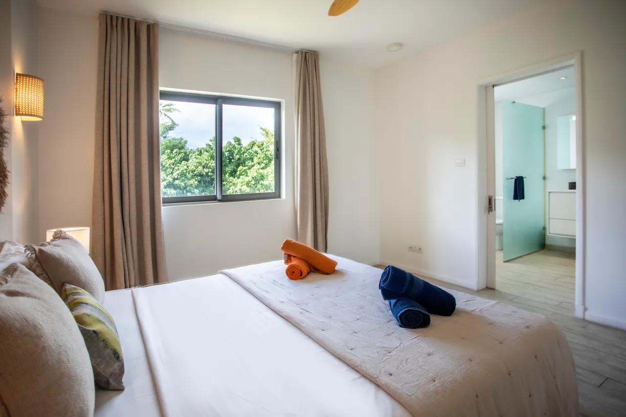 Chambre avec lit double et vue sur le jardin verdoyant.  
Éclairage mural créant une ambiance douce et accueillante.  
Linge de lit coloré ajoutant une touche de vie à l'espace.  
Sol en carrelage clair pour un entretien facile au quotidien.  
Rideaux élégants encadrant la grande fenêtre pour plus d'intimité.