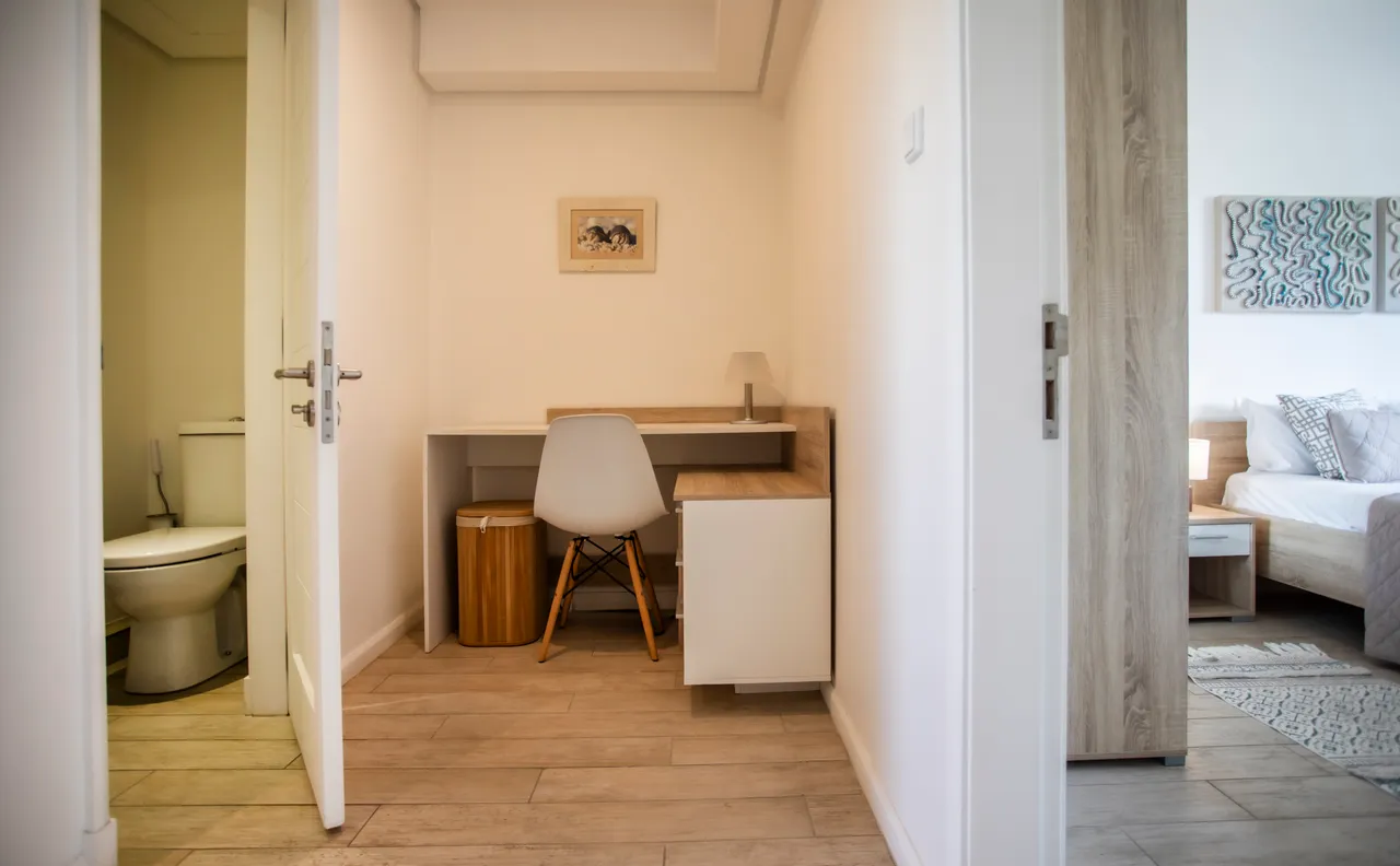 Espace de bureau avec un meuble de travail intégré et une chaise moderne.  
Lumière naturelle filtrant par la porte ouverte créant une ambiance lumineuse.  
Sol en carrelage clair facilitant l'entretien de l'espace de travail.  
Rangement discret avec panier en bois et armoire intégrée.  
Décoration murale sobre ajoutant une touche contemporaine à la pièce.